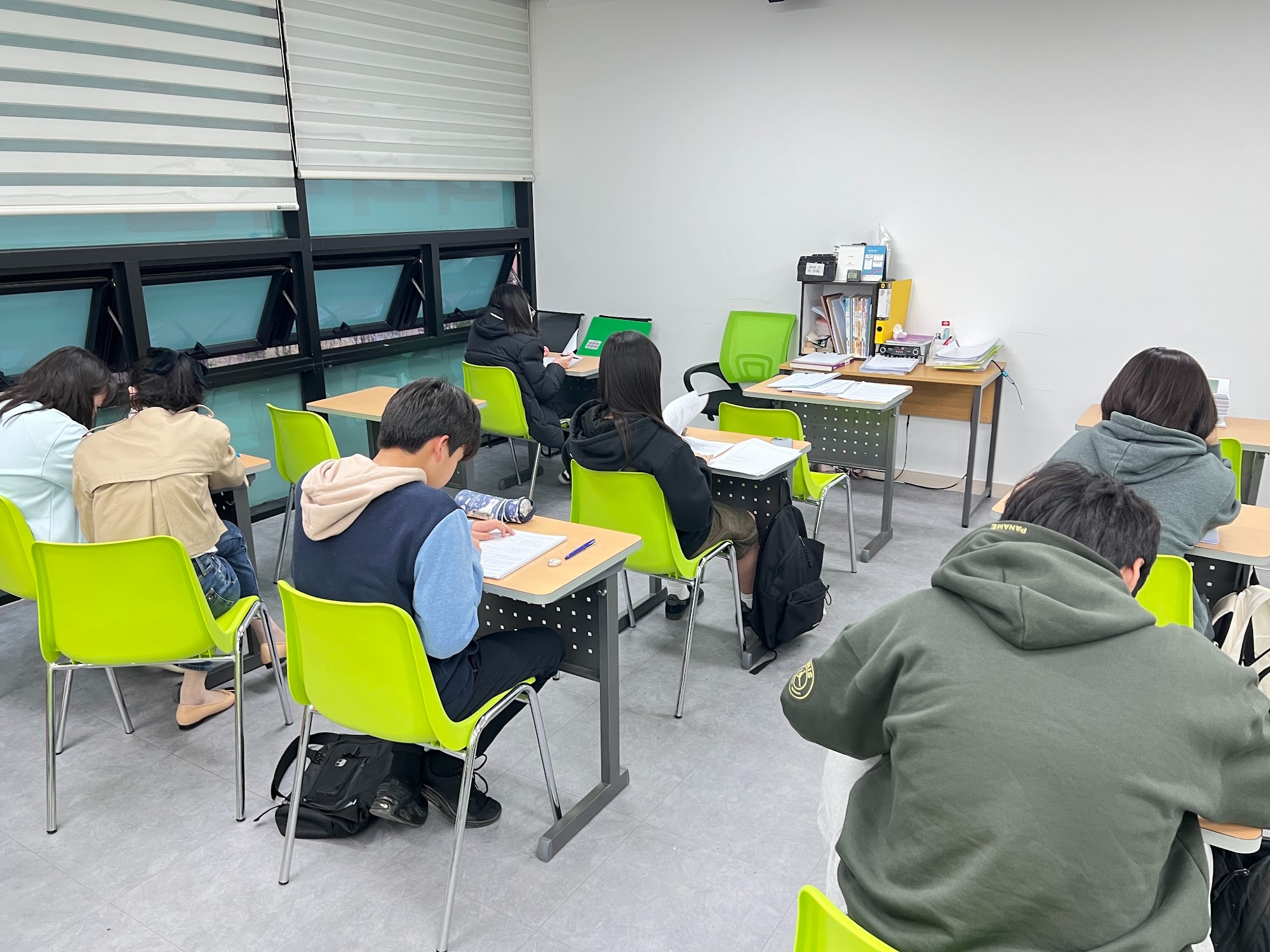 다산지금점 자습 시간 — 학생들이 뒷모습으로 연두색 의자에 앉아 각자 책상 위 학습지를 풀고 있고, 교실 안쪽 코치 책상에는 파일과 자료가 쌓여 있다