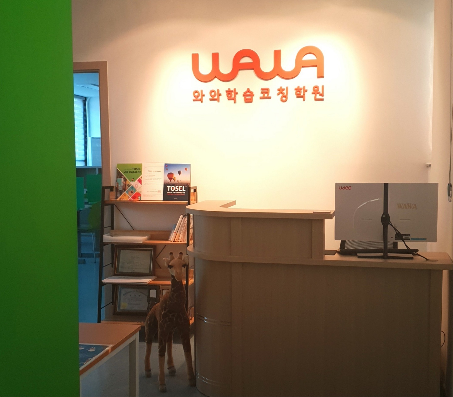 단대점 프런트 — 벽에 주황색 WAWA 로고와 와와학습코칭학원 글자가 붙어 있고, 그 아래 안내 데스크와 모니터가 놓여 있다. 왼쪽 선반에는 영어 시험 교재 안내 책자와 액자가 세워져 있고, 데스크 옆으로 교실로 이어지는 문이 열려 있다