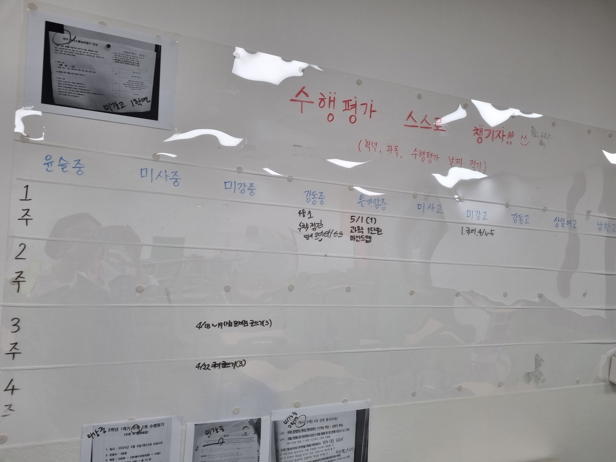 미사점 벽에 붙은 수행평가 일정판 — 흰 벽에 가로줄이 그어져 있고 맨 위에 빨간 글씨로 수행평가 스스로 챙기자라고 적혀 있다. 세로로는 학교 이름이, 가로로는 1주·2주·3주·4주가 칸으로 나뉘어 있으며 칸마다 과목과 날짜가 손글씨로 적혀 있다