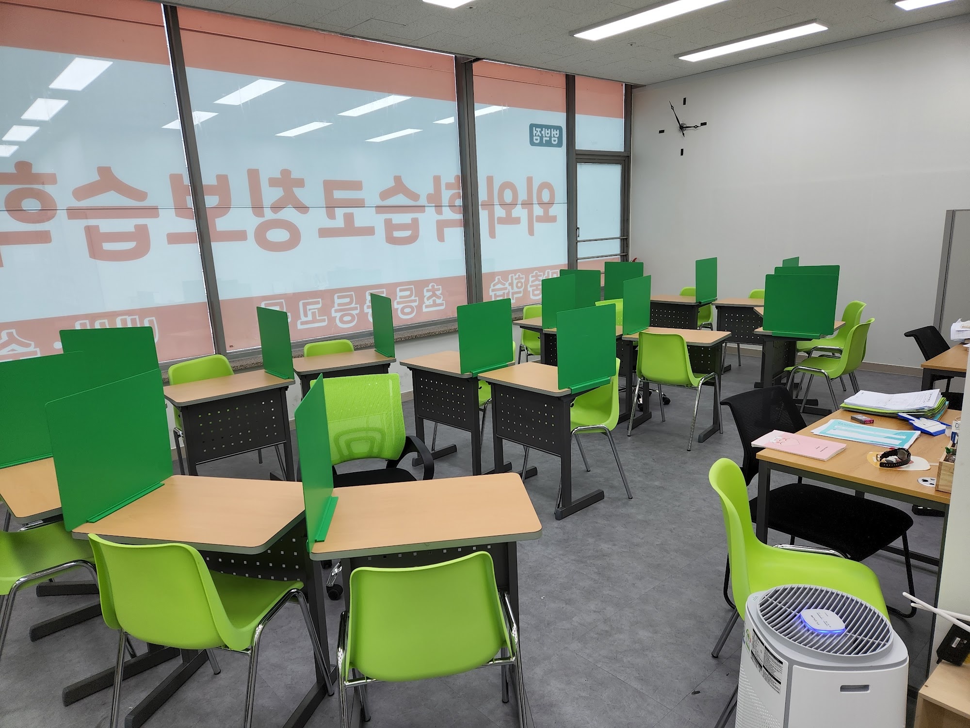 범박점 창가 교실 — 큰 유리창에 와와학습코칭학원 범박점이라고 적힌 시트지가 안쪽에서 비쳐 보이고, 창 앞으로 초록색 칸막이를 세운 1인용 책상이 여러 줄로 놓여 있다. 오른쪽 벽에는 검은색 원형 시계가 걸려 있고, 안쪽에 교재를 쌓아 둔 책상과 공기청정기가 있다