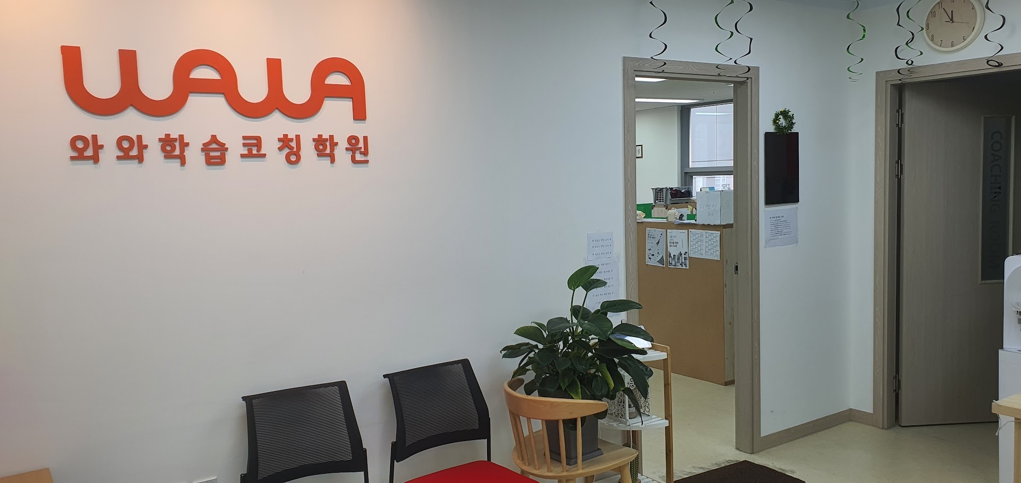 산내점 입구 안쪽 공간 — 흰 벽에 주황색 와와 로고와 '와와학습코칭학원' 글자가 붙어 있고, 그 아래 검은 의자 두 개와 화분이 놓여 있다. 오른쪽으로 상담실과 데스크로 통하는 문이 열려 있고 벽시계가 걸려 있다