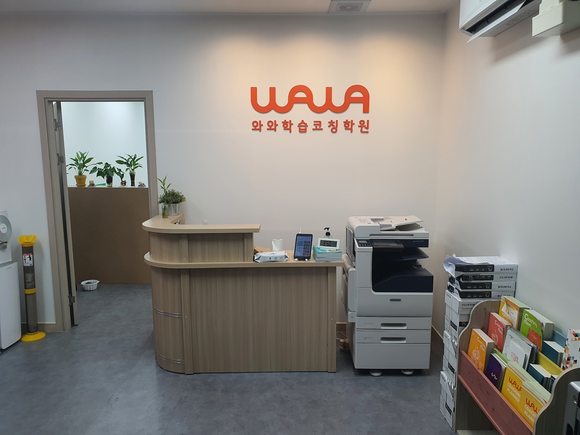 창원 진해구 석동 와와학습코칭 석동점 안내 데스크 — 흰 벽에 붙은 WAWA 와와학습코칭학원 사인, 나무색 데스크와 복합기, 오른쪽에 코칭플래너·코칭맘 책자가 꽂힌 책꽂이