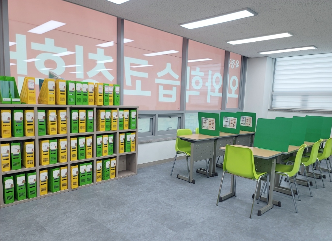양주 옥정동 와와학습코칭 옥정점 교실 — 왼쪽 벽면에 초록색과 노란색 파일박스가 칸칸이 꽂힌 큰 수납장이 있고 박스마다 이름표가 붙어 있다. 뒤쪽 유리벽에는 분홍색 바탕에 '오늘학습코칭'이라고 쓰인 브랜드 월이 보이고, 오른쪽에는 초록 가림판이 세워진 책상과 연두색 의자가 놓여 있다