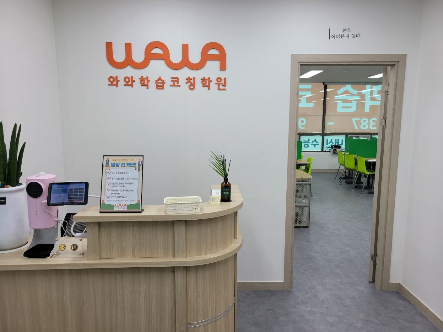 이충점 입구 데스크 — 흰 벽의 주황색 WAWA 와와학습코칭학원 로고, 나무색 곡선 데스크 위 태블릿과 '입장 전 체크!' 안내판, 화분, 오른쪽 벽의 '꿈은 어디든지 있다.' 문구와 열린 문 너머로 보이는 교실