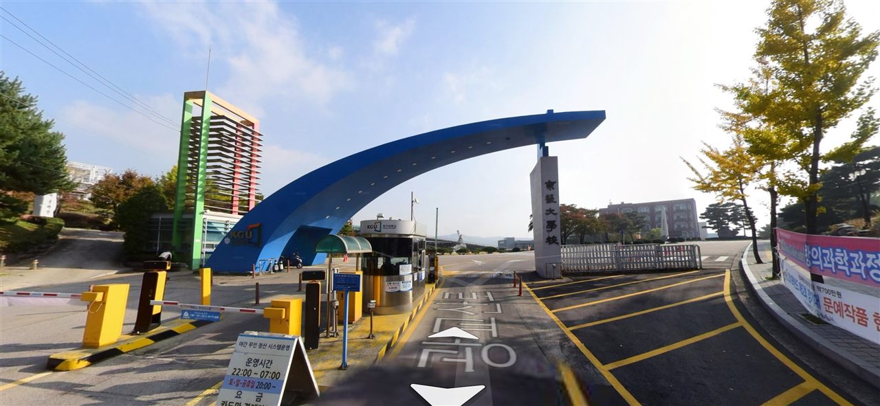 경기대학교 수원캠퍼스 정문(서문) — 넓은 도로 위로 캠퍼스 입구가 이어지고, 길 양옆으로 나무와 캠퍼스 건물이 보인다