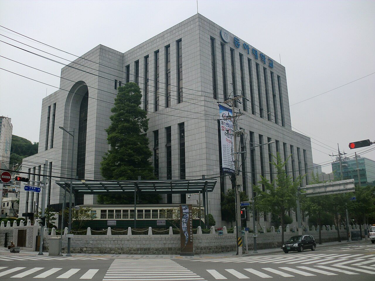 동아대학교 부민캠퍼스 — 교차로 모퉁이에 선 회색 석재 외벽의 각진 건물로, 위쪽에 동아대학교 교표와 파란 글자 간판이 붙어 있다. 건물 앞 낮은 돌담 안쪽에는 유리 지붕을 씌운 옛 전차 한 량이 전시돼 있고, 앞쪽으로 횡단보도와 신호등이 보인다