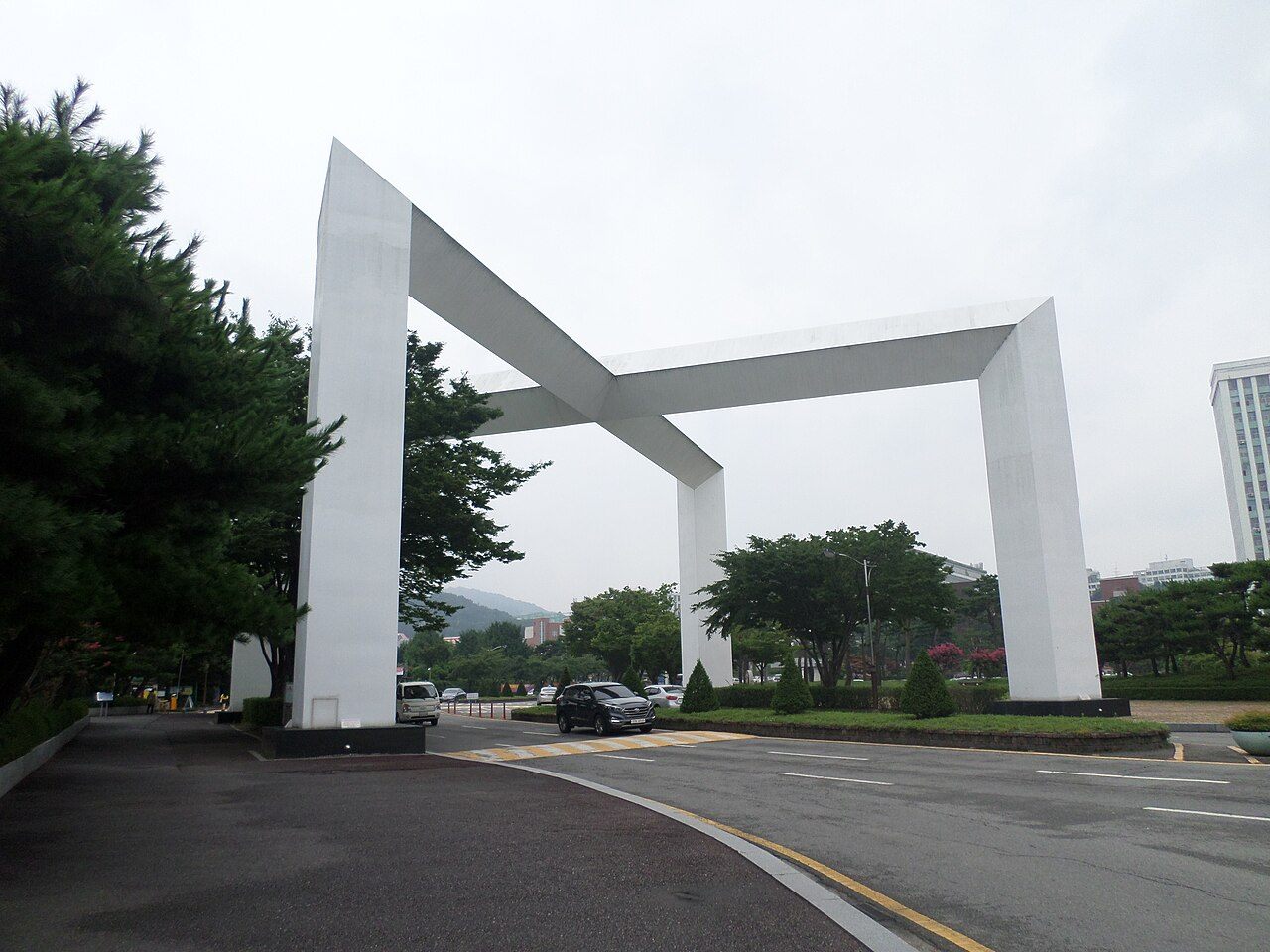 조선대학교 정문 — 흰색 콘크리트 기둥 두 개가 비스듬한 보로 엇갈리게 이어진 대형 조형 문과 그 아래를 지나는 차도, 좌우의 소나무와 잔디